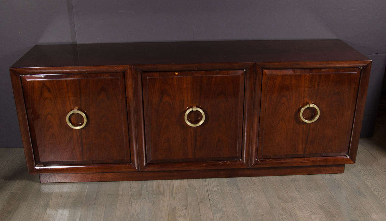 This sophisticated Mid-Century Sideboard by  T.H. Robsjohn-Gibbings for Widdicomb features three cabinets with centralized brass ring pulls and is finished in ebonized mahogany. Each cabinet has an adjustable shelf and the interior of the cabinet