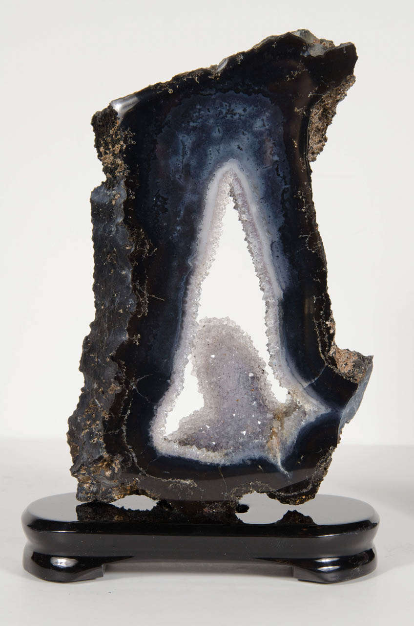 This sliced geode specimen is mounted on a custom made ebonized walnut base. It features an angular form in dark tones ranging from black, grey & deep blue and has a centre encrusted with sparkling quartz crystals.