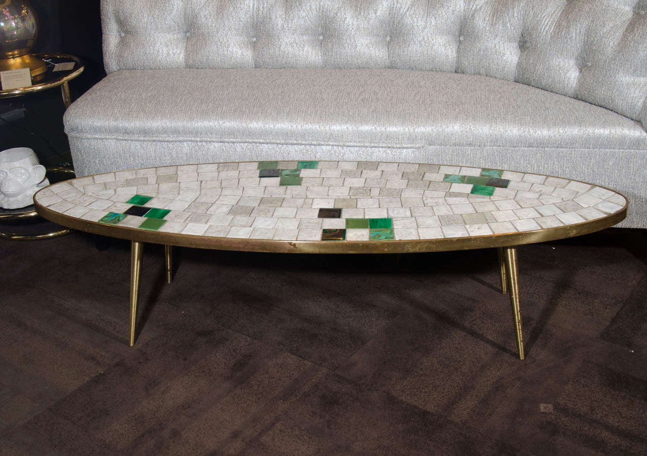 Modernist cocktail table with elliptical form. Comprised of a tiled top with mosaic patterns and a heavy-weight patinated brass base with tapered legs.  The tiles are primarily in hues of white, grey, and ivory with random tiles in various shades of
