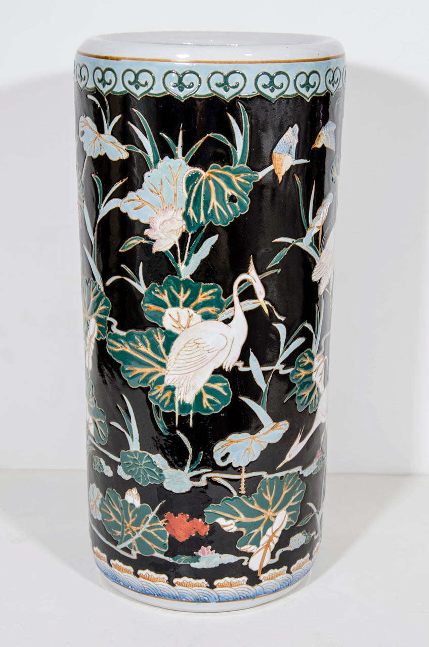 Glazed porcelain pottery umbrella stand or large decorative vase with hand painted Oriental designs.  Has cylindrical form and features a variety of Heron birds and flowering lily pads in vibrant colors over a black backdrop, all of which have been