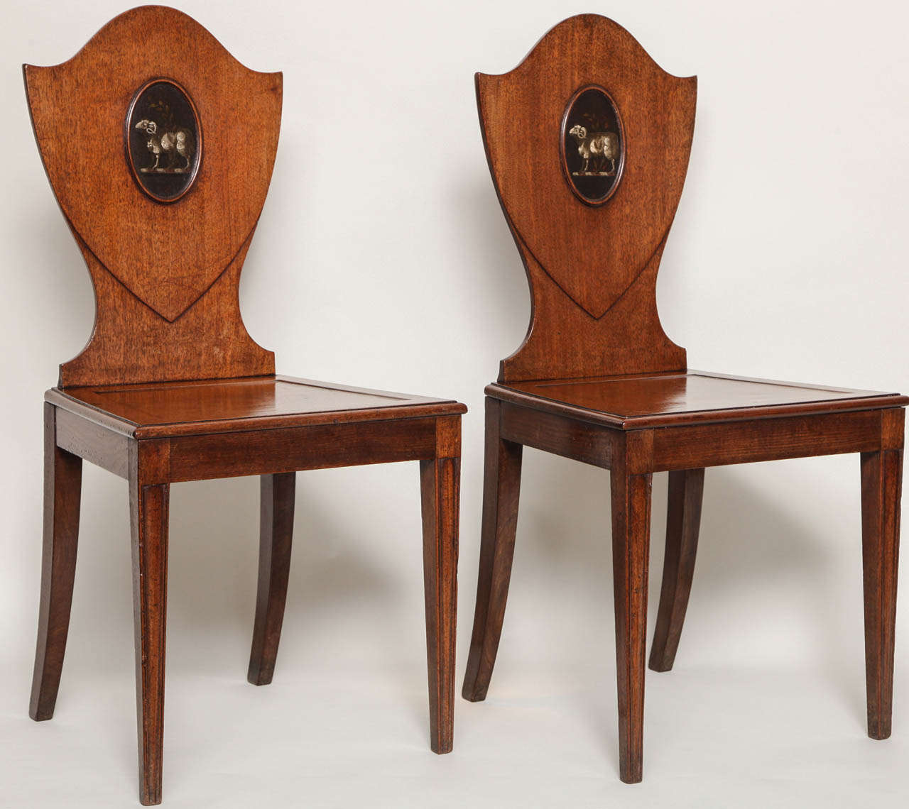 Fine pair of George III mahogany hall chairs, attributed to Gillows, the shield backs with oval crests having heraldic painted rams with laurel branches, the seats with square recess and molded edge, standing on molded tapered legs, both with good