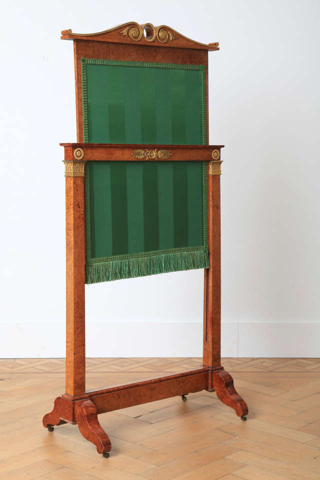 This fire screen has a scrolled top above an adjustable rectangular sliding panel of framed green fabric, on square tapering supports, united by a stretcher on downswept legs, terminating in castors. The gilt bronze ornaments show floral