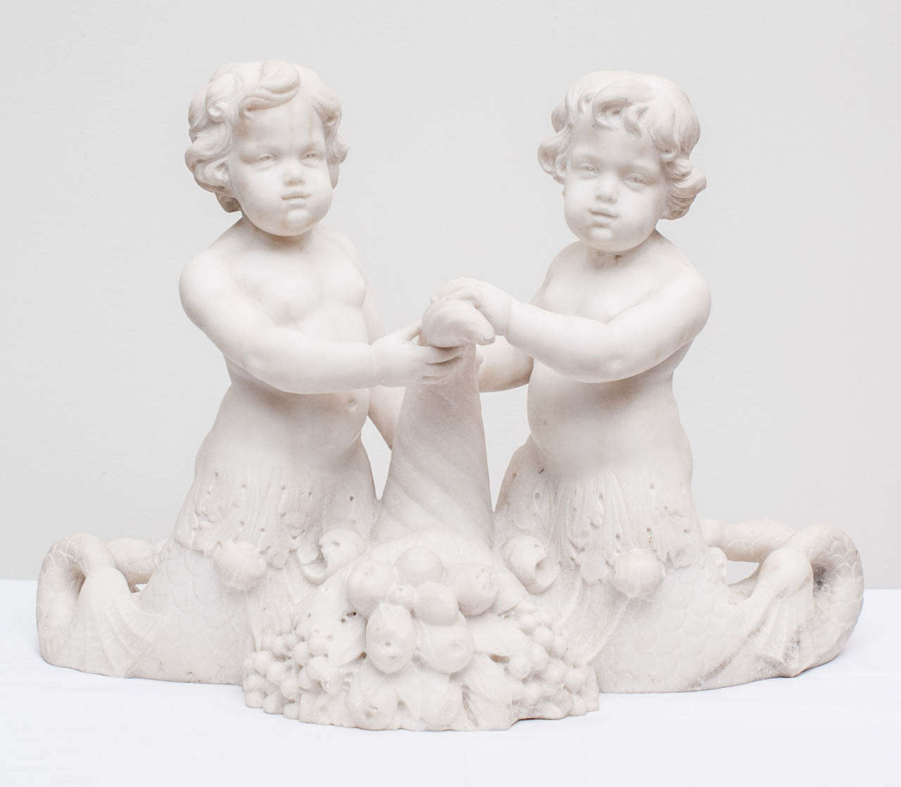 A  carved Carrera marble figural group depicting dolphin-tailed cherubs flanking a cornucopia, circa 1920.