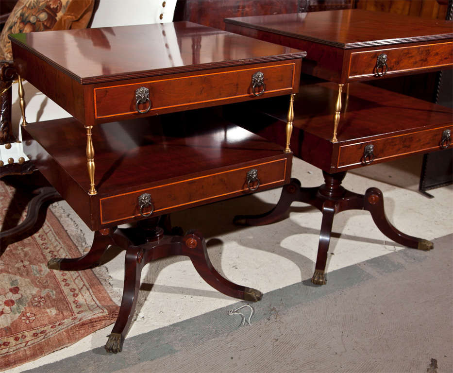 Pair of English Regency style end tables labeled Schmieg & Kotzian, Inc., each has two-tier with single drawer supported by brass uprights, raised on a single pedestal with splayed legs.