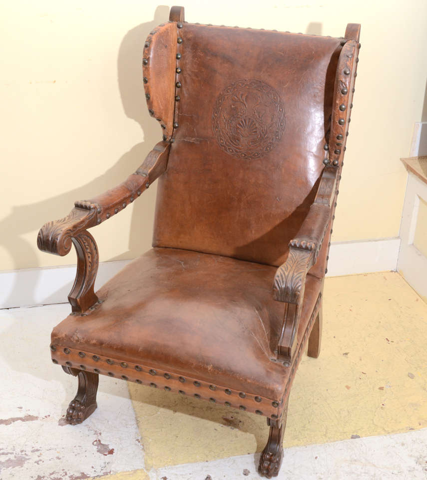 DUTCH LEATHER UPHOLSTERED LATE 19TH EARLY 20TH CENT. 0PEN ARM WING CHAIR WITH CARVED SCROLLED ARMRESTS & LION PAW FRONT FEET. BACK EMBOSSED WITH CIRCULAR WREATH CENTERED WITH A SHELL FLANKED BY 2 DOLPHINS. LEATHER ORIGINAL WITH LARGE NAILHEADS