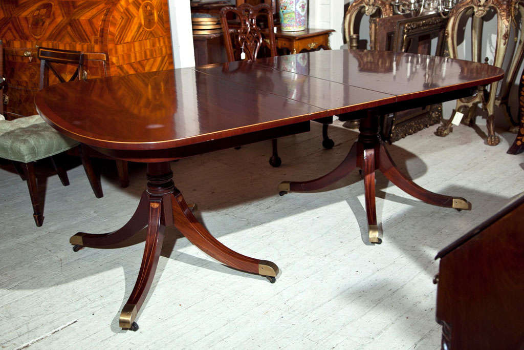 A double pedestal dining table in mahogany with cross banding in mahogany and ebony and satinwood stringing, Each pedestal with 4 legs, splaying from a  central column, each leg with string inlays in a V form, topped by an inlaid fan. Brass cap ends
