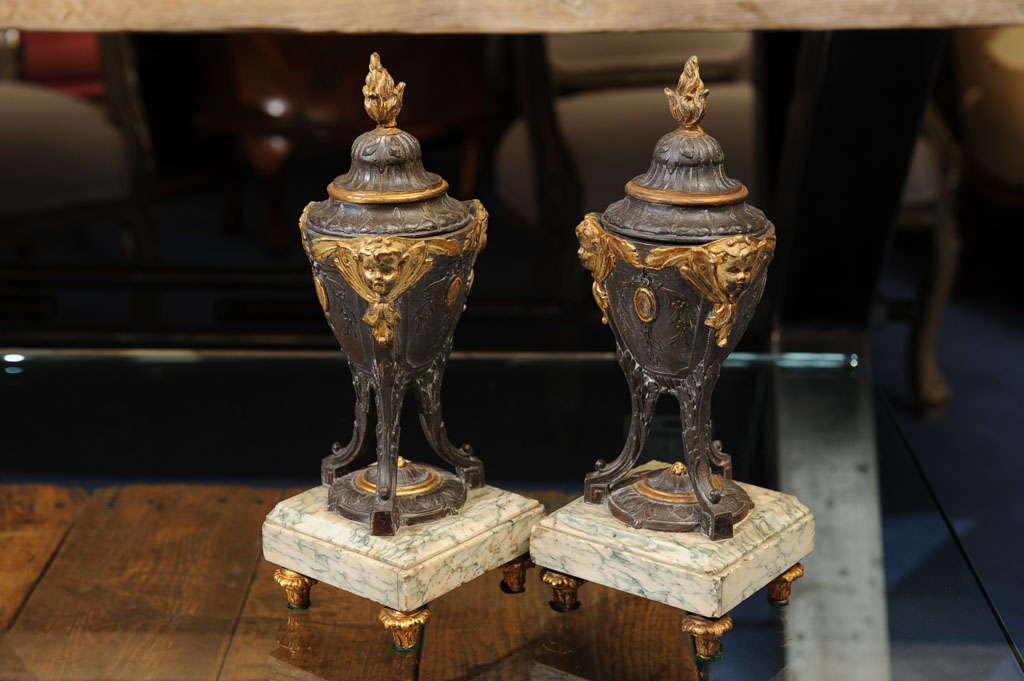 Each cast urn decorated with gilt putti raised on three supports above stepped marble plinths ending on toupie feet, each covered with low dome lids surmounted by flame finals.