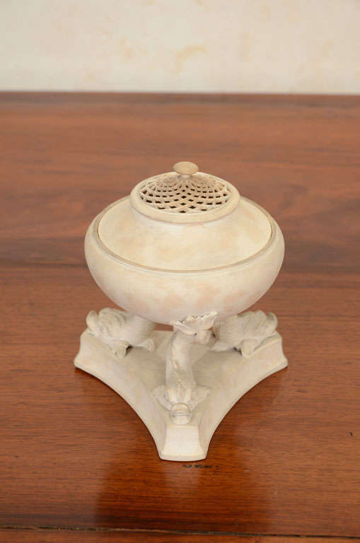 This fine English pastille or incense burner, made at the Wedgwood manufactory, has a reticulated lid resting on a rounded body raised on dolphin-form supports on a concave-sided, triangular plinth, all in a matte, buff-colored ceramic. Based