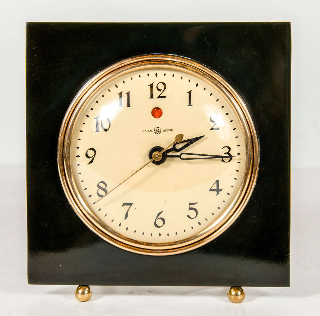 Art Deco Streamlined Vitrolite and brass clock by General Electric.  This streamlined Art Deco clock features a square surround made of Vitrolite that frames the clocks round brass outlined face and rests atop two stylized geometric spherical feet