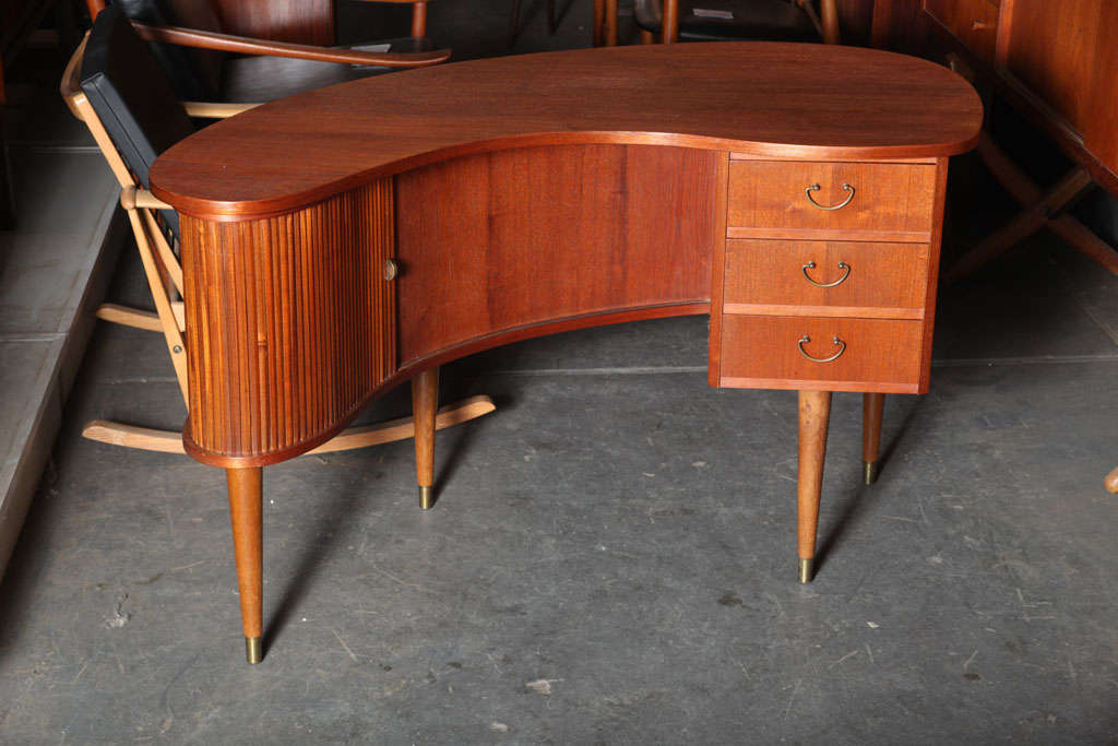 Teak Atomic Era Kidney-Shaped Desk with Tambour Doors.  Features  brass feet and hardware, and a boomerang-shaped interior bar with a glass shelf.  The front side of the desk houses open shelves and an additional tambour door which opens to reveal a