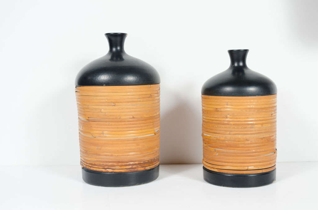 A pair of matte black ceramic jugs comprising small bud vase like necks, cylindrical body wrapped in strips of cane.  Distributed by Raymor.  Italian, circa 1950.