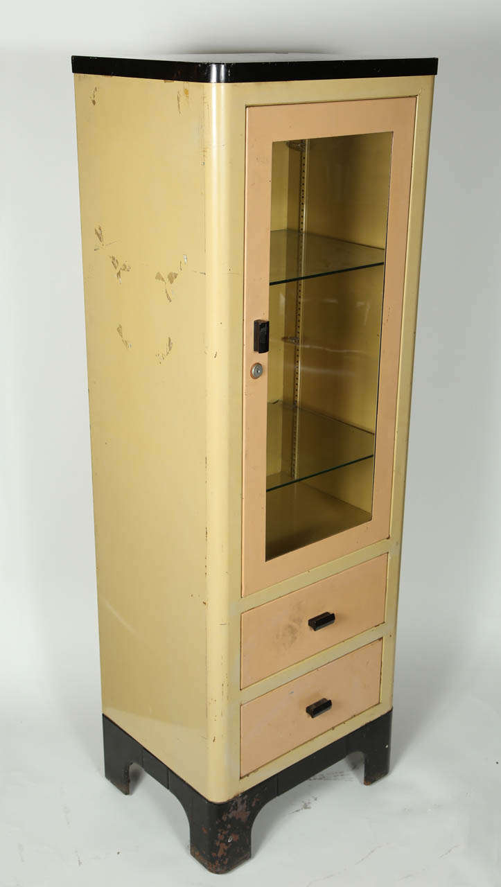 This is a painted metal free standing vintage medical cabinet. This cabinet is painted three different colors in its original state. The two drawers and glass door are painted a peach color while the cabinet itself is painted a tan. The feet and top