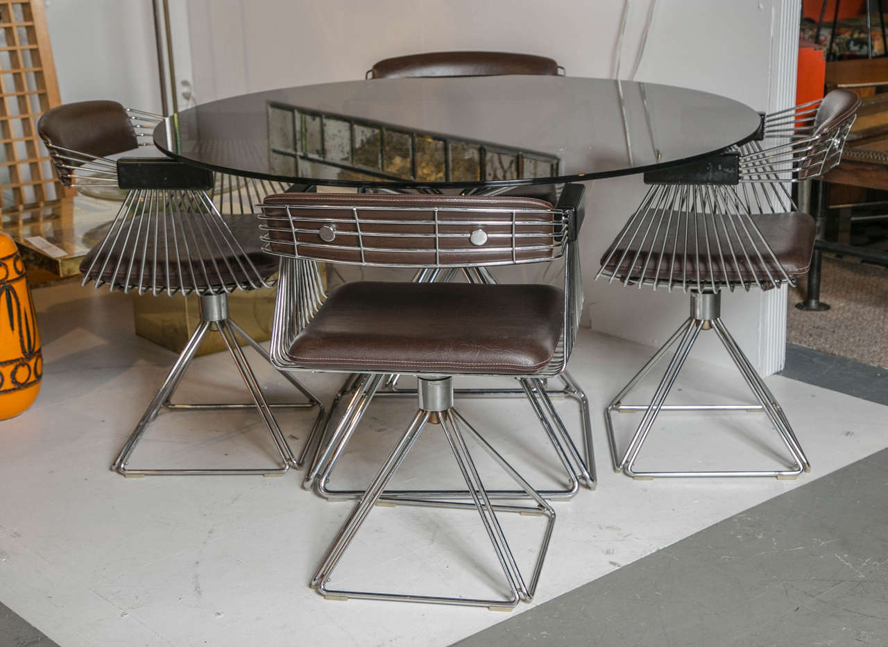 A Rudi Verelst dining set. A round smoked glass and pyramid chrome base dining table, with matching swivel armchairs of chrome and original brown vinyl and black lacquered armrests.