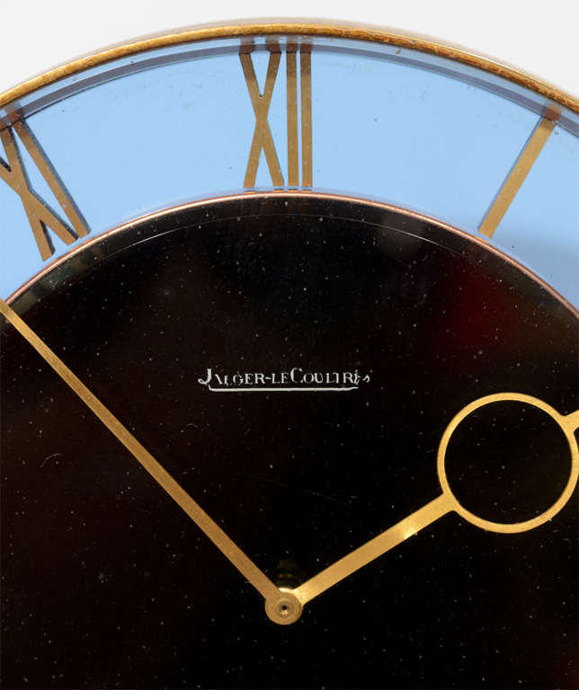A vintage Art Deco tabletop clock by renowned Swiss watchmaker Jaeger-LeCoultre. the piece has a round black dial with Roman numerals set against a blue glass ring.