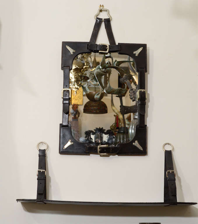 A vintage French mirror and wall mounted shelf, each with saddle leather and nickel silver detailing. The set is in the style of Jacques Adnet. The piece is in good vintage condition with age appropriate wear; leather stitching loose in some