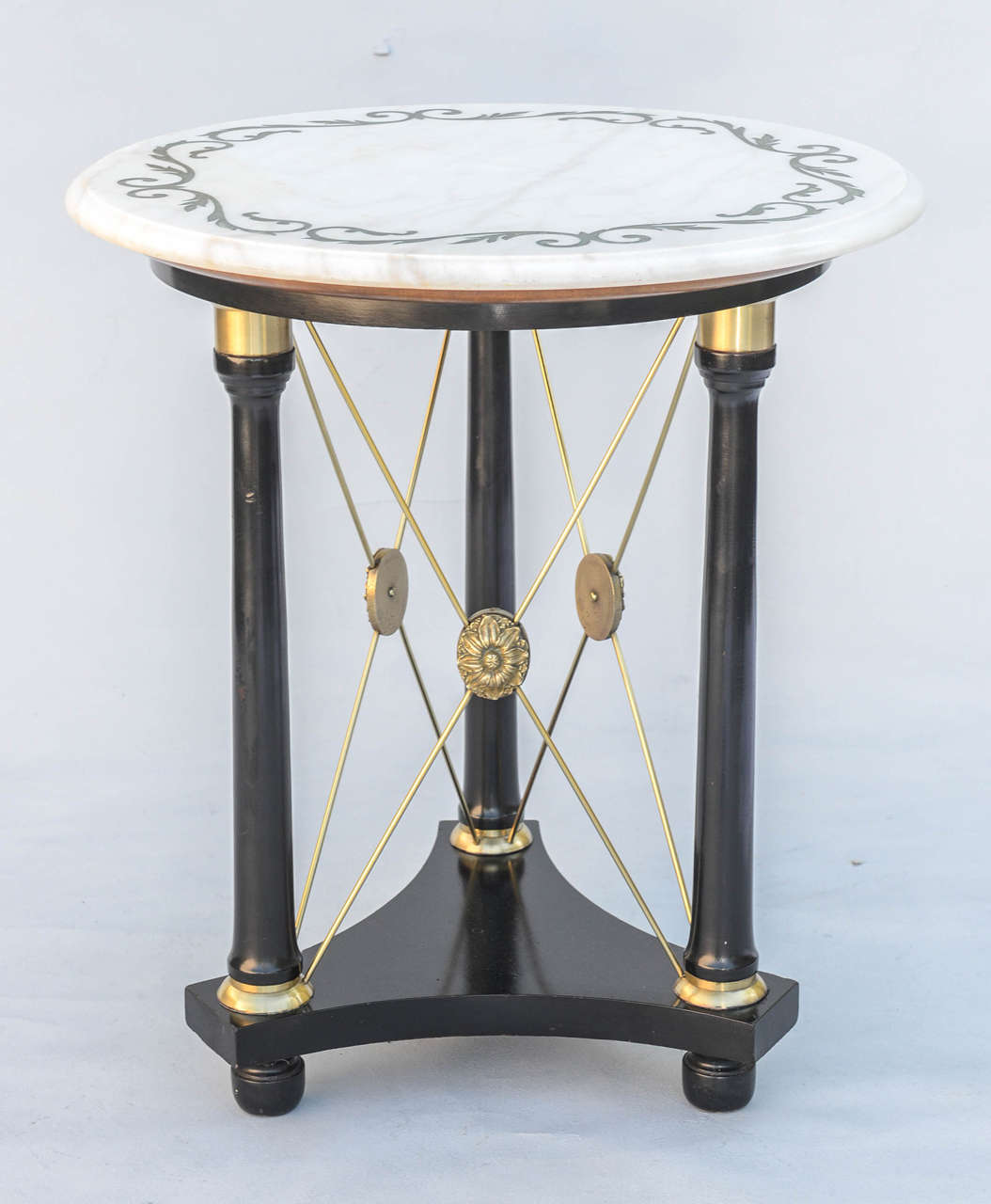 Unusual occasional or end table, in Regency taste, having a lacquered finish; its round top of Carrara marble etched with scrollwork border, on a trio of brass-capped column legs, joined by X-frame crossed brass bars, each centered with a stylized