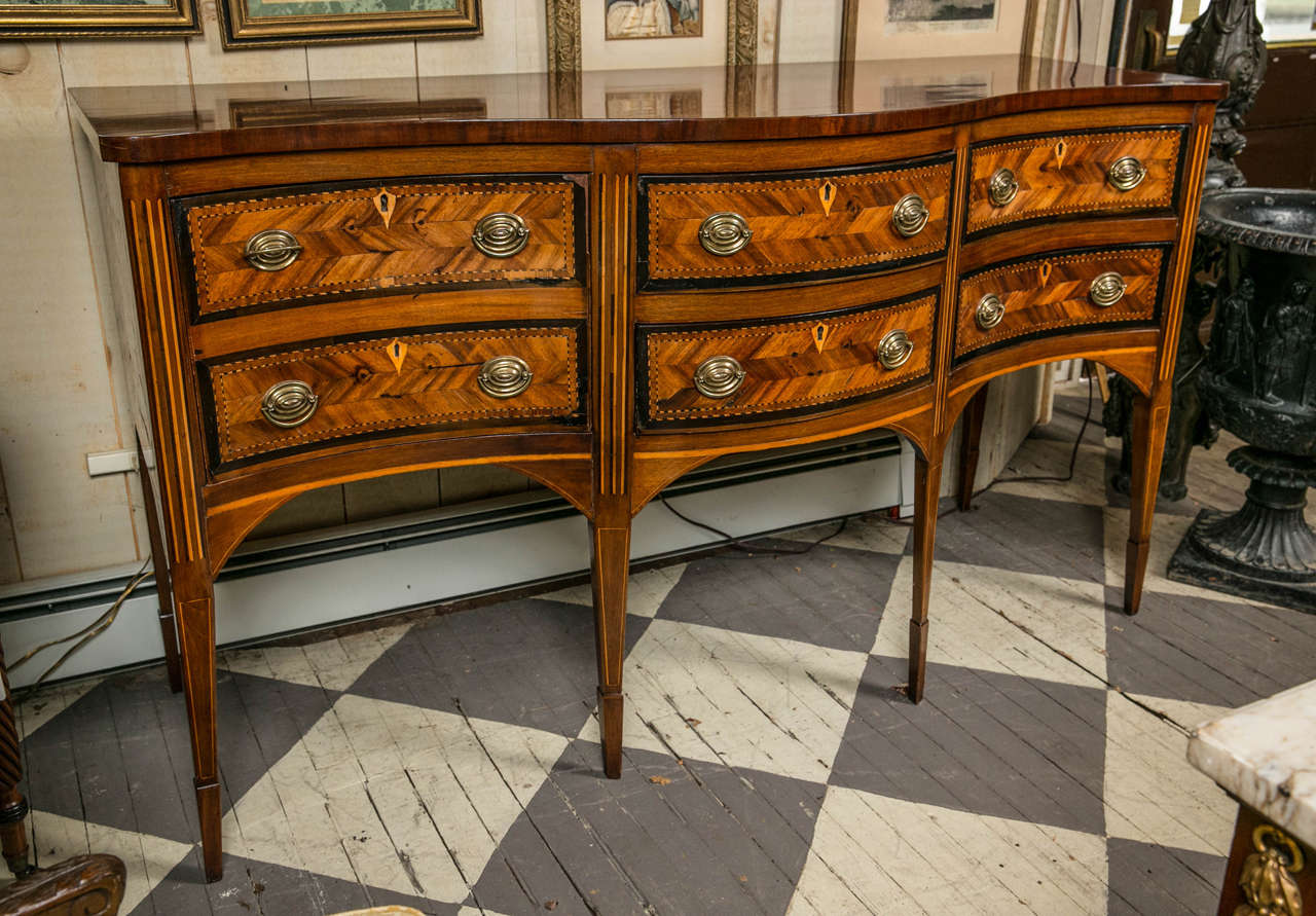 Made in the late 19th, early 20th century, this sideboard of mahogany with ebony or ebonized wood and satinwood inlays, has all the bells and whistles of the antique. Graceful S-curved front, bottle drawer to the left, two drawers in the center and
