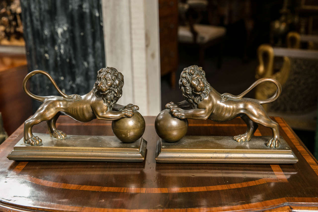 Each lion, one facing left, one right, have finely detailed faces and bodies.
Their front paws rest on a large orb. Their tails swoop up and end on their backs. They are mounted to a three sided bronze base. There are holes at the corners of each