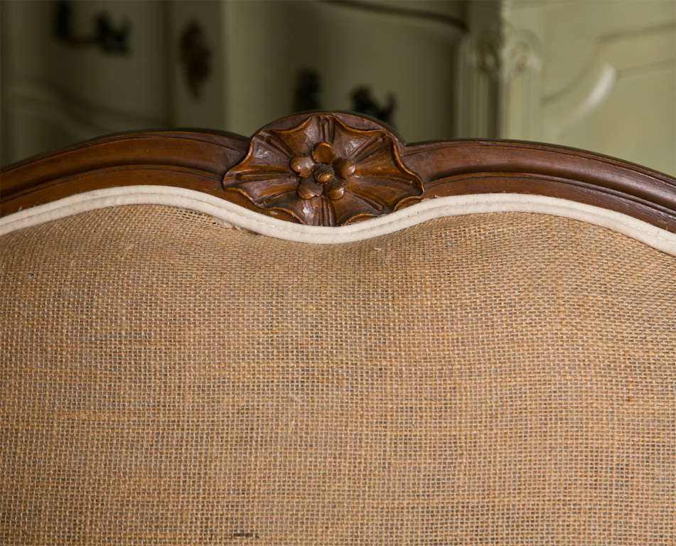 Pair of French Provincial style walnut settees, circa 1950s, each upholstered in burlap and linen, shaped back with padded arm and cushioned seat, raised on short cabriole legs. 