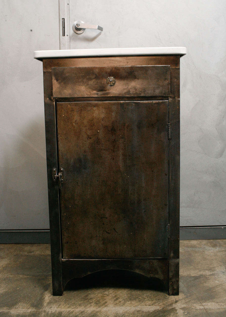 This  compact metal cabinet is utilitarian and elegant with a nicely shaped base and a great enamel top. The metal body of the cabinet has been polished and waxed to acquire a irridescent sheen. This piece has one drawer with a glass handle, a