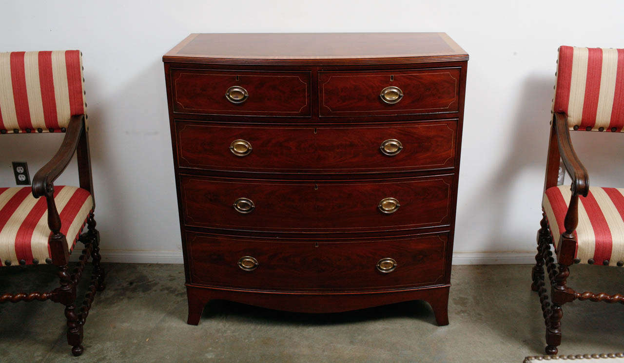 This lovely bow front chest of drawers has satin cross banding, readed top edge and string inlays throughout. The secondary wood is English White Pine. It has graduated, 2 over three drawers. The chest has brass: drop handles, escutcheons and locks.