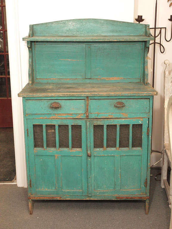 Rustic painted wooden cupboard with high shaped back and narrow open upper shelf.  The back, paneled below the shelf, is framed on either side by carved wooden brackets.  The one on the left has a crack in the upper part.  Two drawers with rusted