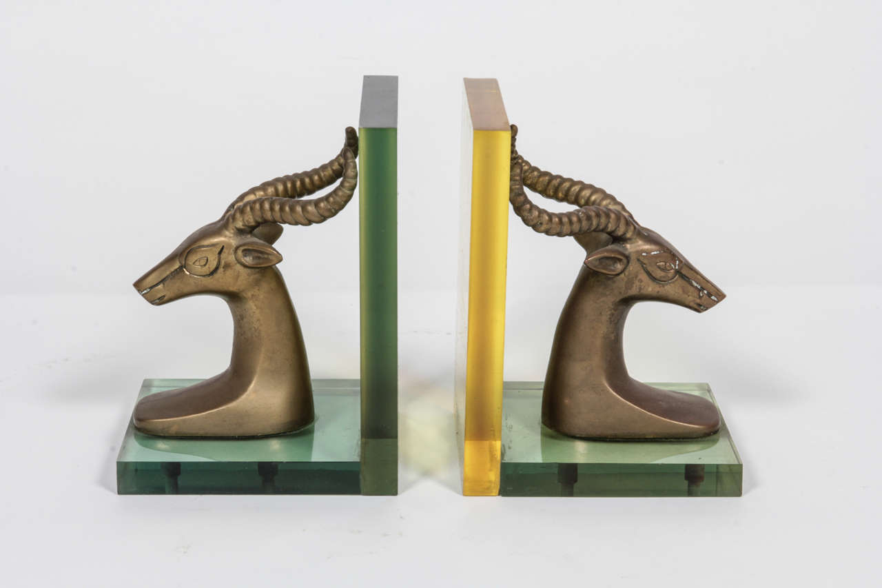 Pair of ram head (Ibex, antelope) bookends in the style of Tony Duquette.  USA, circa 1960.

Metal forms in antiqued brass finish mounted on Lucite bases; one bookend has a green-hued Lucite base; the other a yellow hue.  Beautifully made.