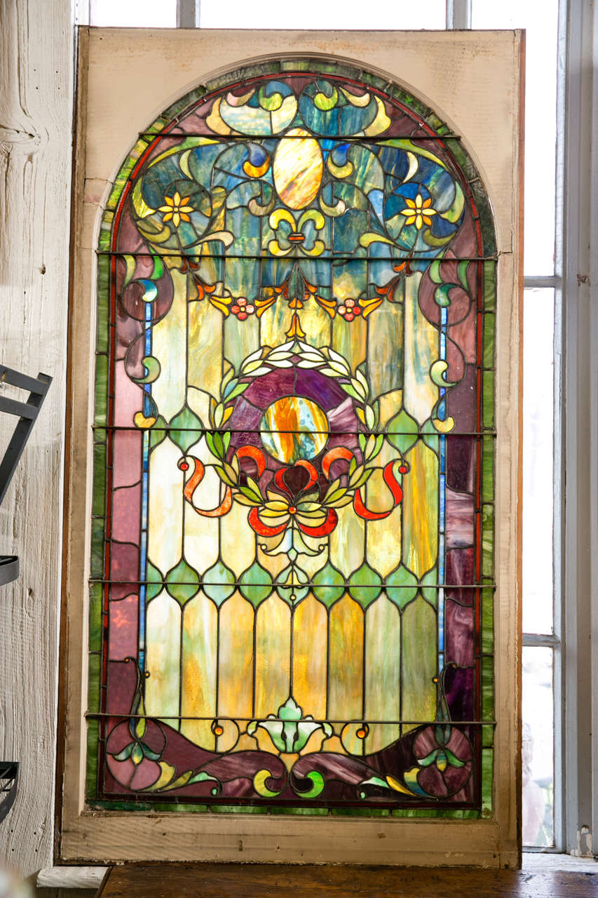 This large antique stained glass window features 13 jewels and an arched top with vibrant color. It has a wreath design in the center. This is one of six matching which were salvaged from the stairwell of the Buena Vista Estate in Greenwich, CT,
