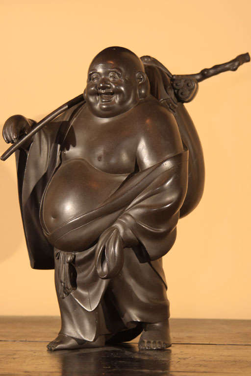Japanese cast bronze figure of Hotei, the god of happiness and prosperity. Hotei, with his characteristic large earlobes and belly, is a symbol of wisdom; dressed in a robe, his treasure bag suspended from a gnarled branch slung over his shoulder