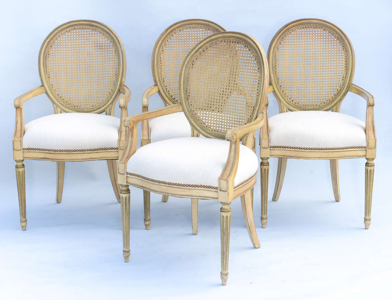 Set of four fauteuils, with painted fielded frames, each rounded caned back flanked by bowed arms, on down-swept terminals, crown seat of linen with nailheads, raised on round tapering fluted front legs, ending in touipe feet, with rear sabre