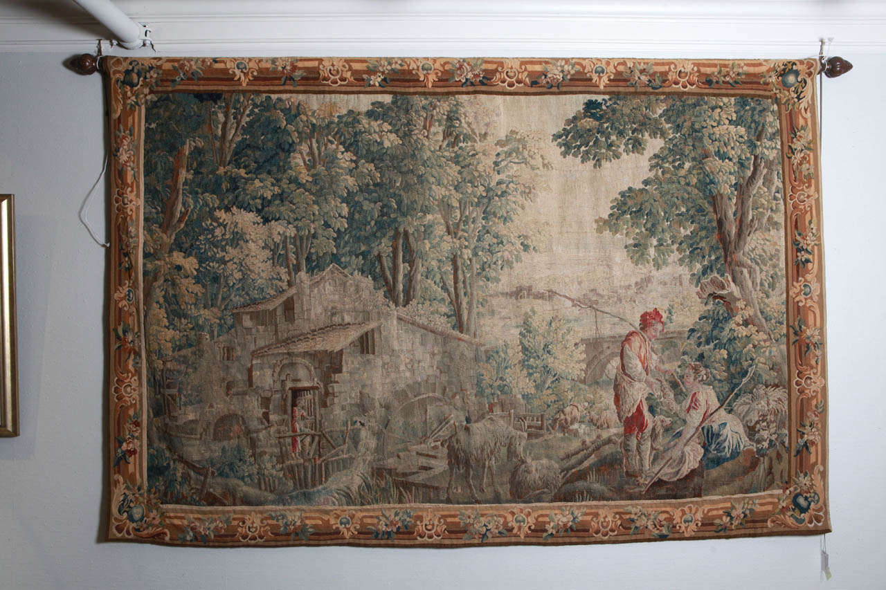 The French 17th century tapestry is telling a charming story of a young woman being courted by a gentleman suitor while a mother awaits anxiously from the
mill house. The rich colors of the wooded area in greens and browns are predominant with a
