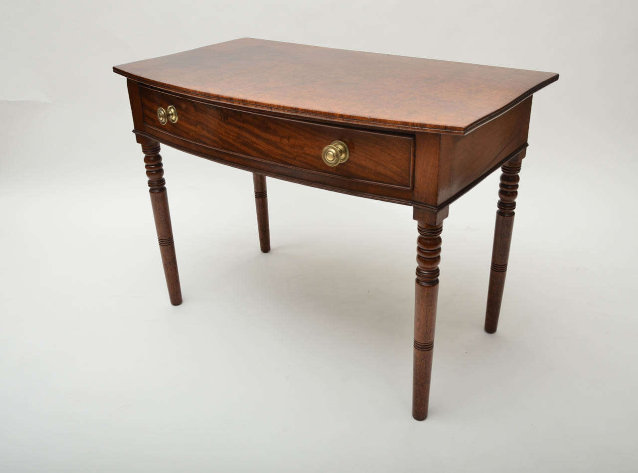 An early 19th century Regency mahogany bow front side table with one drawer, turned legs, and brass ring pulls.