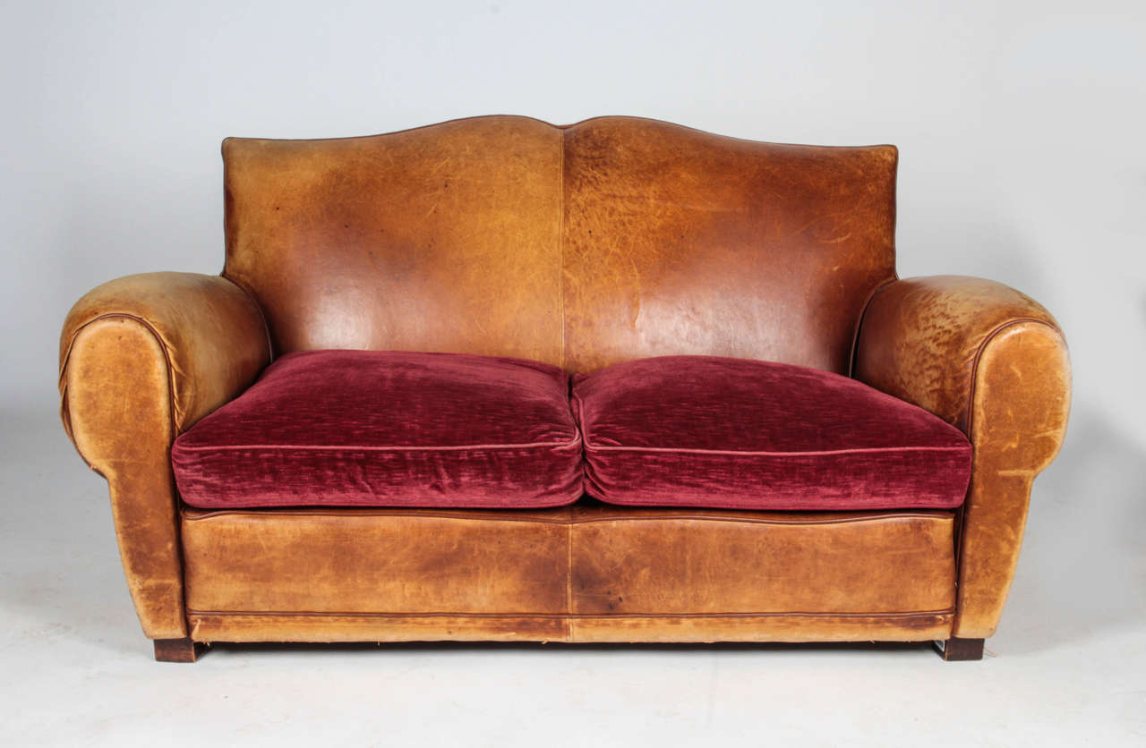 This tan leather loveseat has original leather that was altered to look aged and worn. Chocolate-colored leather piping contrast and accentuate the curvatures of the arms and mustache-style back. Cognac-colored chenille seat cushions create a