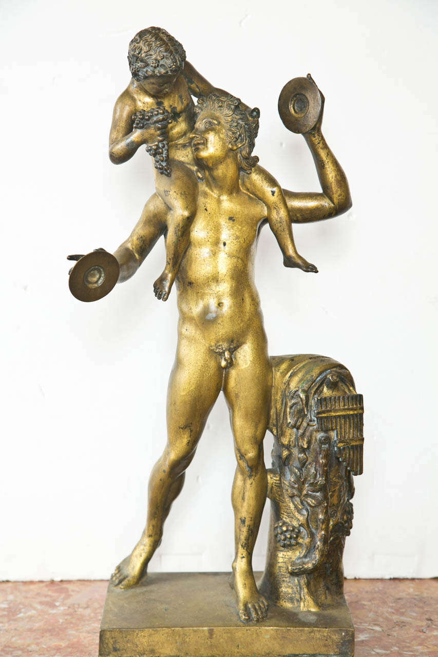 The  gilt bronze, nude  figure of Bacchus stands next to a  tree trunk form draped with a cloak , grapes and a set of  pan pipes.. He hold on his shoulder a putto who offers to the god, a  bunch of grapes. In his hands he holds a pair of