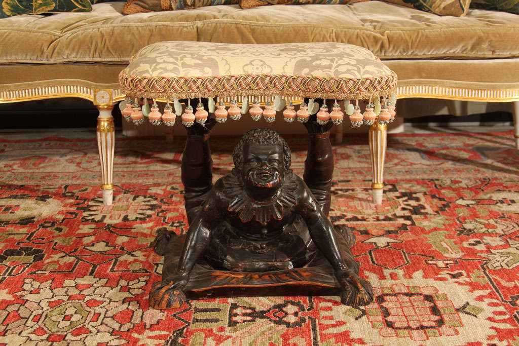 A 19th century  Venetian carved wood Blackamoor Stool depicting an acrobat on a cushion. The original seat is upholstered in a vintage piece of Fortuny fabric wile the trim is from Janet Yonaty.  A similar painted version of this stool may be seen