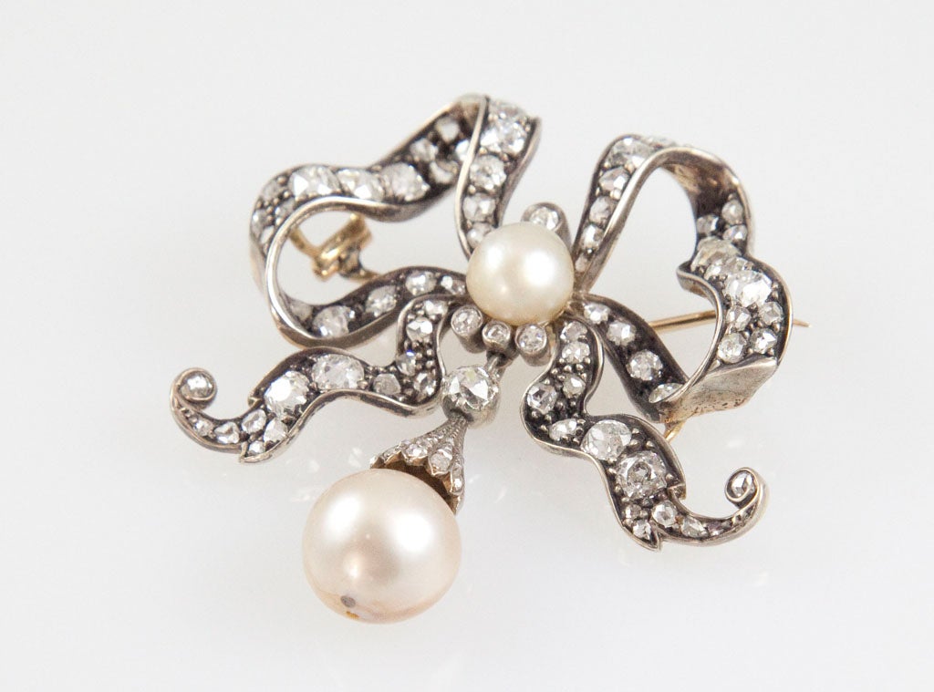 backed in gold, topped in silver and bead and bezel-set with diamonds. The center of the bow is set with 1 pearl and suspended from the bottom is 1 natural pearl in a diamond cap. The pearl measures 12.46 x 11.30 x 11.05 mm and is accompanied by a