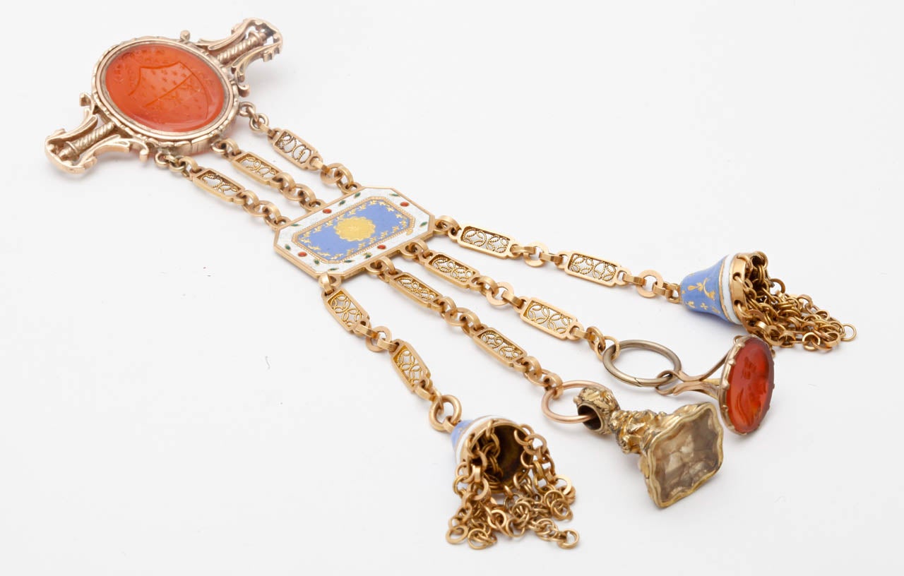 A Carnelian, enamel and gold chatelaine. The oval carnelian plaque is carved to depict a family crest, suspending 3 fancy link chains and an octagonal enameled plaque in shades of blue, white red and green, terminating in 4 fancy link chains.

All