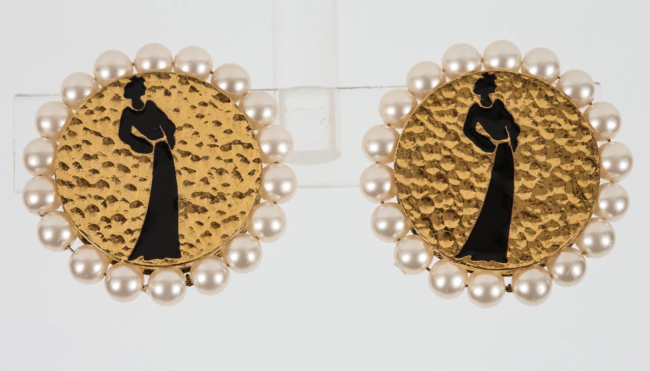 A lovely pair of hammered gilt metal ear clips by Chanel with an enameled silhouette of Coco in a long skirt and a frame of glass pearls. The backs of the earrings feature the 