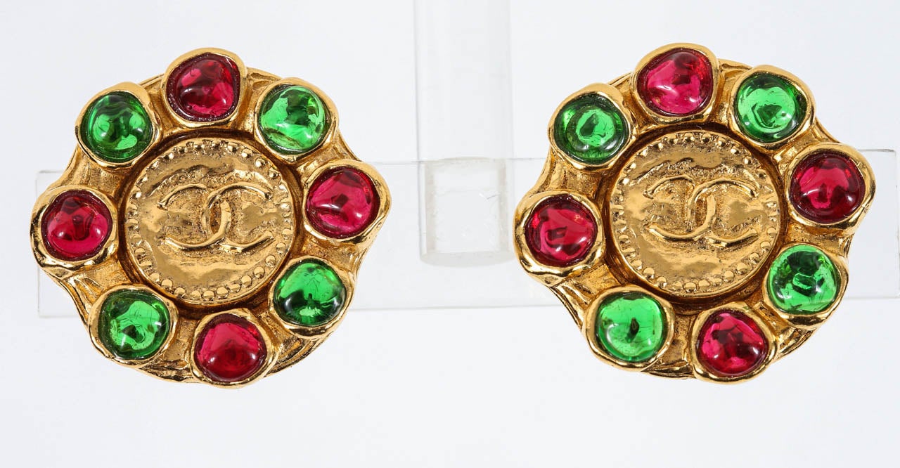 A classic pair of Chanel ear clips with a beaded edge center coin featuring the interlocked C logo, framed by irregular red & green poured glass stones. Marked on the backs of the ear clips with the 