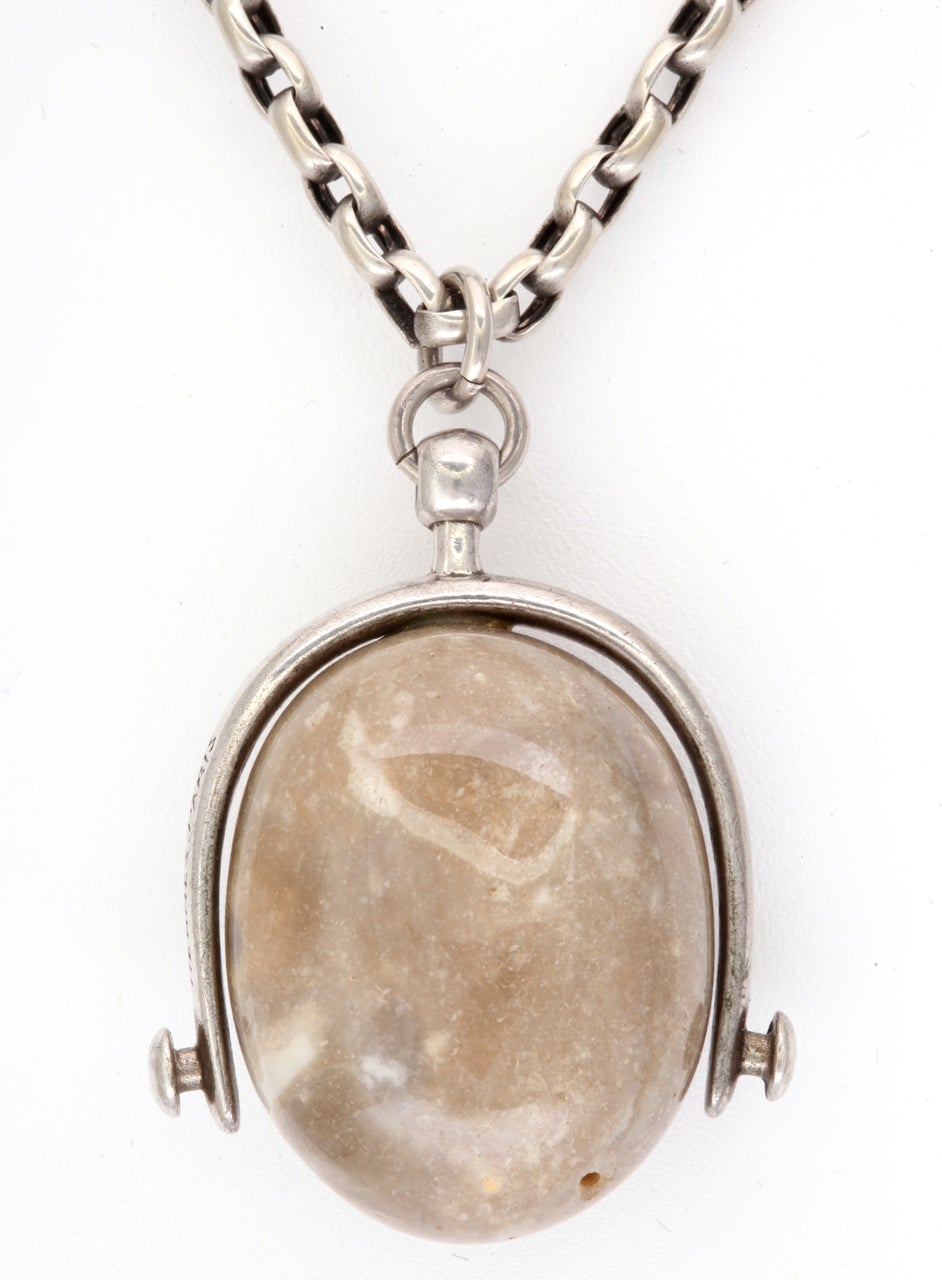 A large equestrian silver stirrup grasps a  palomino stone that has been polished to a pleasurable, tactile smoothness. The stone's natural colors are a blend of beiges, grey and white. The enlargement exaggerates the eccentricities in the stone's