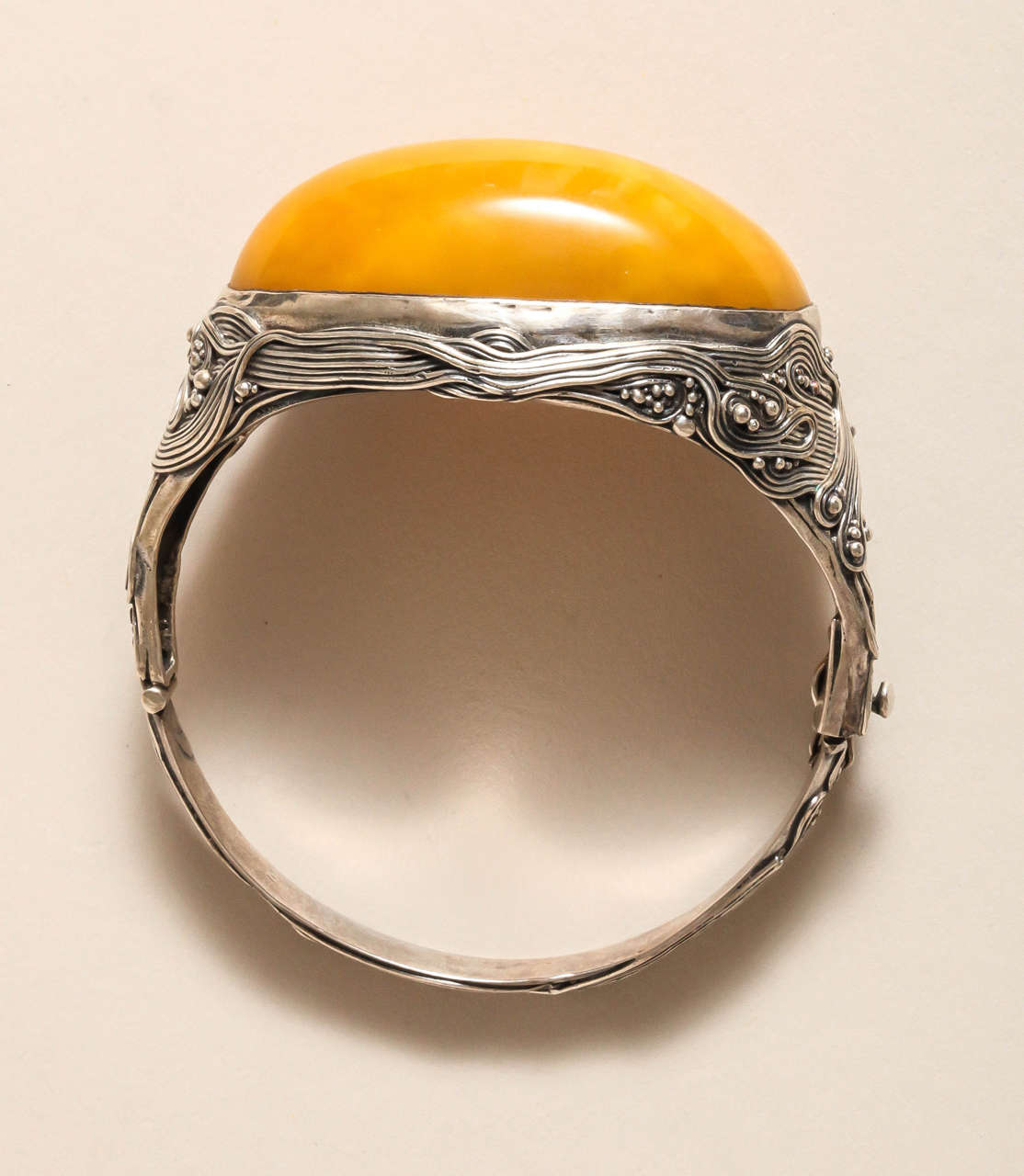 The bangle is set with a large cabochon-shaped amber stone in beautiful warm butterscotch shade.  It is decorated with small intricately entwined lines which run all the way around and are enhanced by tiny berries or pearls.  It has a v-shaped clasp