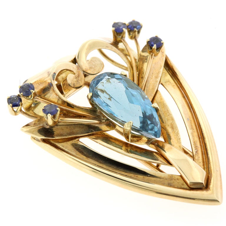 Tiffany & Co circa 1940's arrowhead-shaped clip brooch in 14K gold typifies the Retro Modern style, with its broad curved scrolls at top, and geometric tapered form, set at center with a pear-shape blue topaz and accent blue sapphires. Clip measures