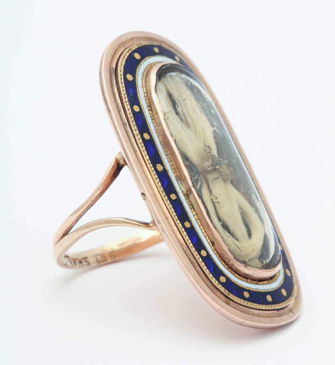 An 18th century ring of spectacular design in an elongated oval form displays a bow of white hair tied with gold thread placed under its crystal. The crystal frame is gold framed by white enamel. Outstanding is the next oval of strong blue enamel