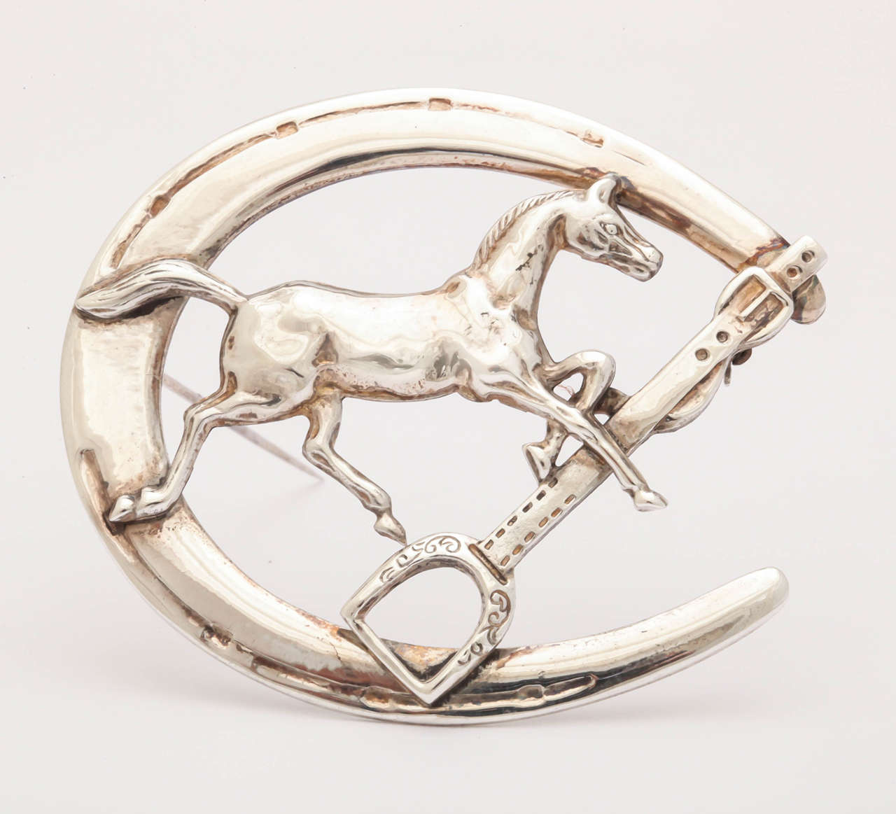 This sterling silver pin is a classic and stunning piece of Equestrian jewelry. It  features a prancing horse , stirrup with leather strap encircled by a horse shoe. The pin is an impressive size, 2 in. by 2 1/2 in.  This design is also available as