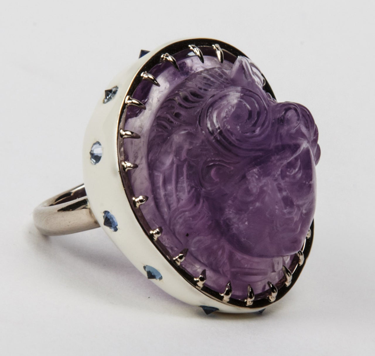 Ring in 18K white gold, partially lacquered in white, set with small sapphires, culet-up, centering an important amethyst carving of a head of Eros, 18th century work in the antique taste.
The carving is an original antique; the mounting is a