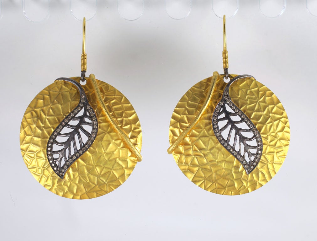 A pair of 18kt yellow gold, rhodium plated sterling silver and diamond earrings. The gold discs have a textured vein pattern and have a gold stem wrapped around the edge of each earring. . There is a rhodium plated sterling silver leaf with diamonds