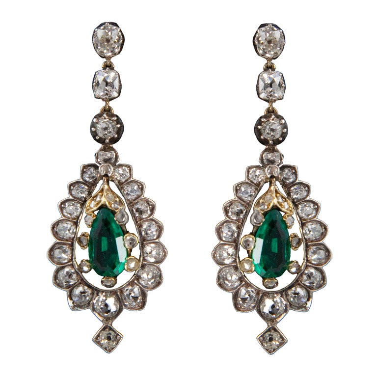 Each earring is backed in 15 karat gold, topped in silver and centers 1 pear-shaped emerald in a six-prong, diamond-set mount surrounded by diamonds in modified scallop mountings, suspended by 3 diamonds in crimped-collet mountings and completed by