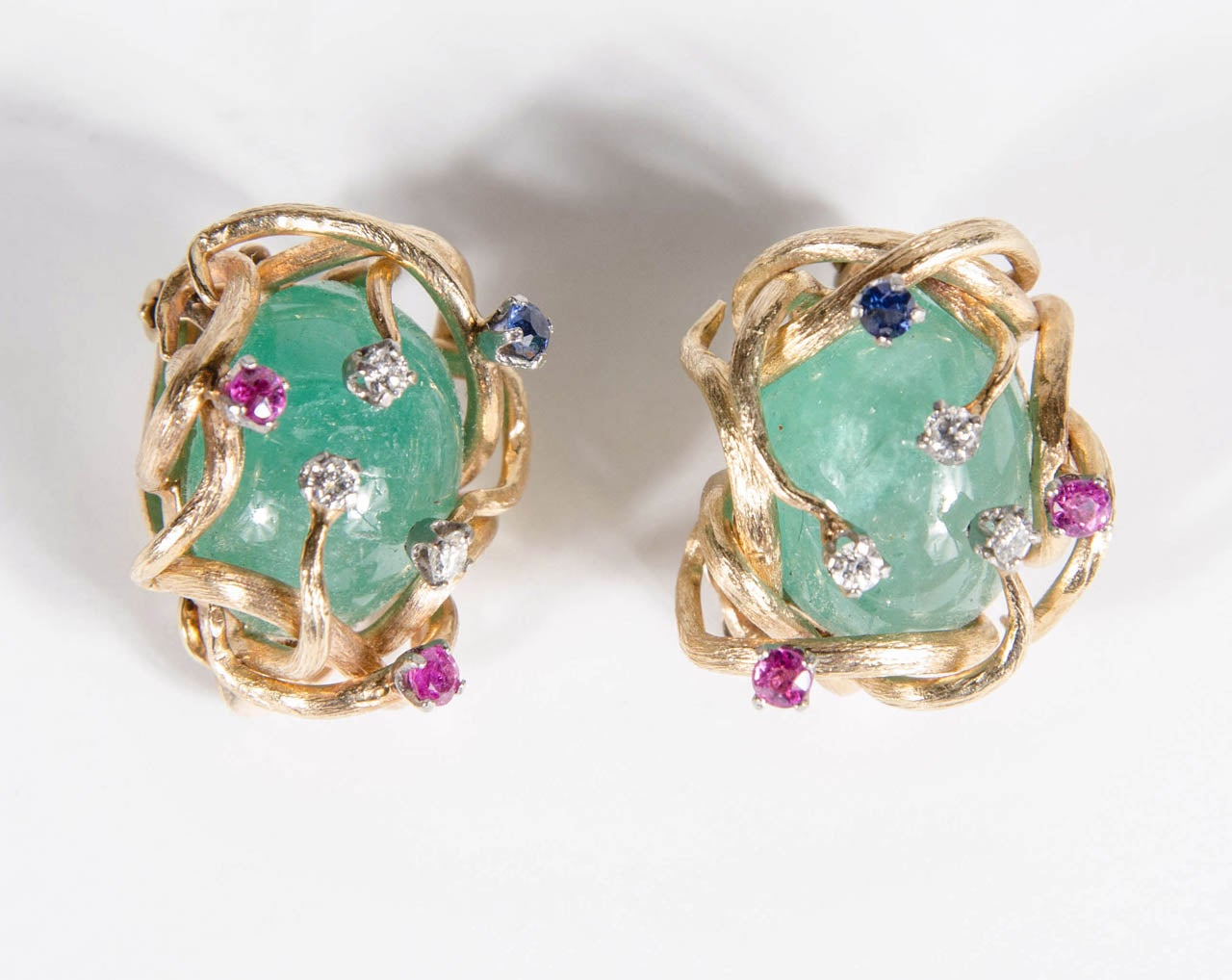 These stunning ear clips feature a cabochon emerald and further mounted with diamonds ,rubies and sapphires. 14.8 dwt inclusive. A great modernist design that features scrolling gold tendrils holding the stones.These can also be converted to pierced