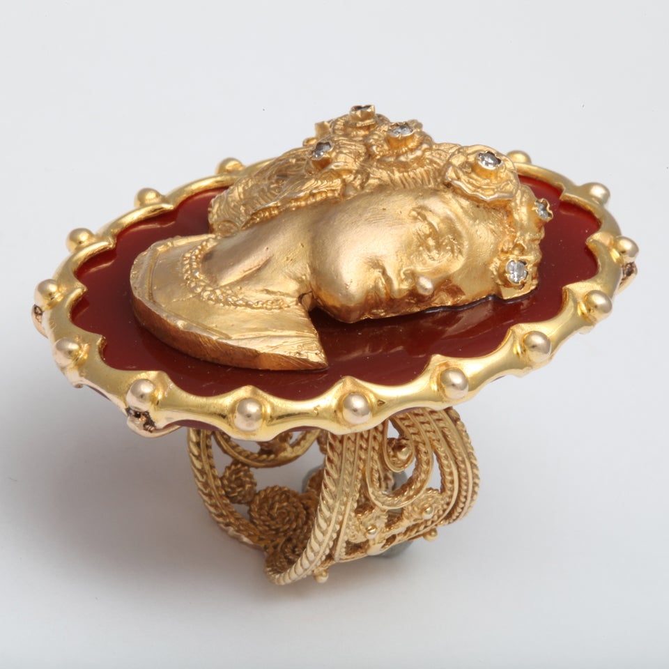 A cameo ring composed of an 18kt yellow gold casting of a cameo, set on an oval agate plaque. The agate is framed with an 18kt yellow gold scalloped border. There are 7 diamonds set in the floral crown. Approximate diamond weight .26ct. size 6