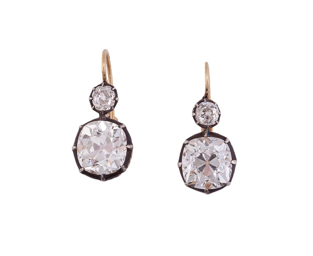 Each earring is backed in gold, topped in silver and centers 1 large old cushion-cut diamond in a crimped collet mounting suspended from 1 smaller old European-cut diamond, also in a crimped collet mounting. Two large diamonds: 1 at 4.80 carats and